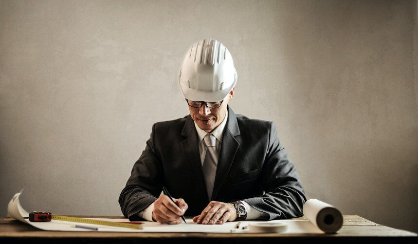 Person in a suit and hard hat writes on papers at a desk with a ruler and blueprint.