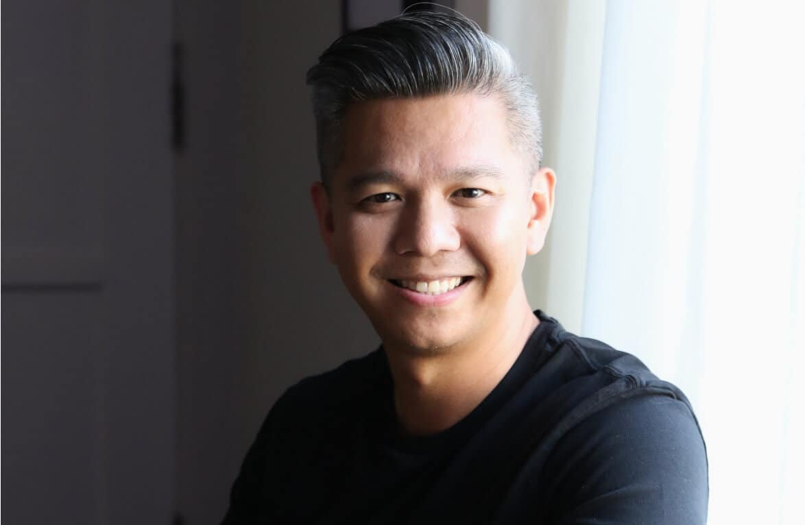 Smiling person with short gray hair in a black shirt, sitting by a window with light curtains.
