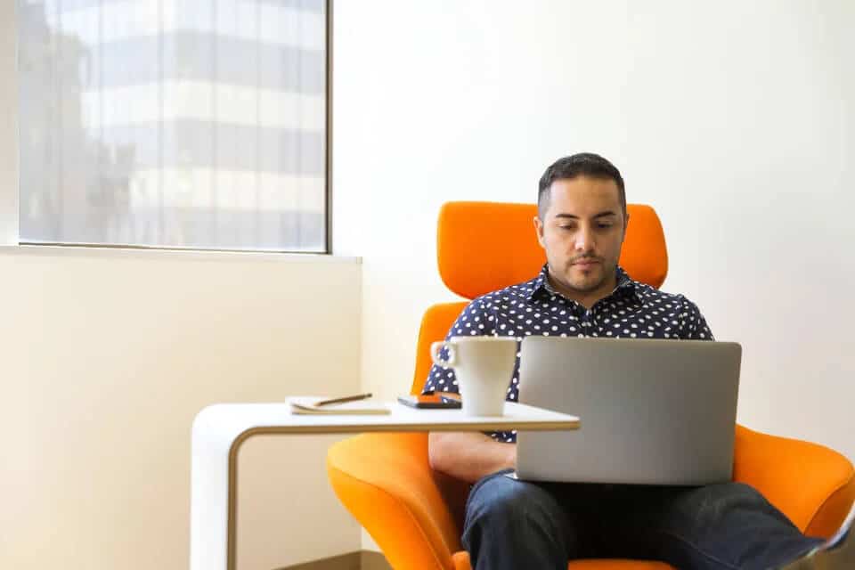 Man seated in an orange chair, using a laptop on his lap, with a table holding a phone and cup nearby.