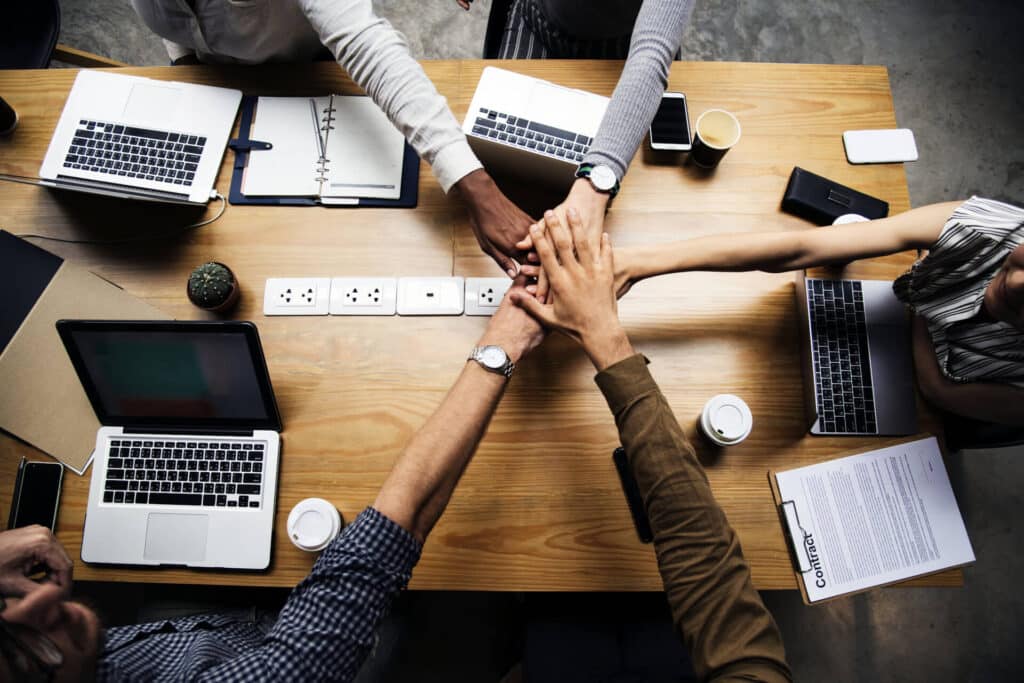 Six people at a wooden table stack hands over laptops and papers, showcasing teamwork and collaboration.