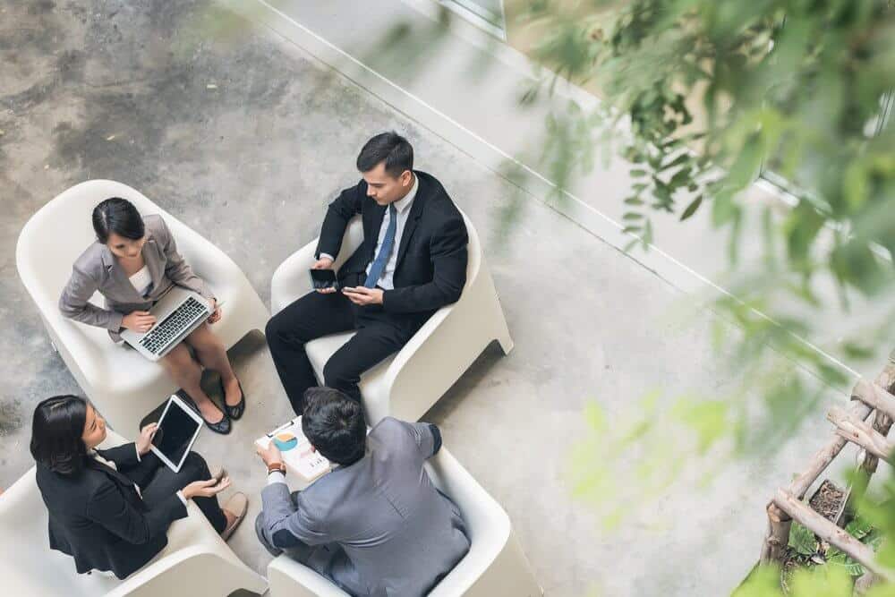 Four professionals in a meeting, sitting in white chairs, discussing over documents and using devices.