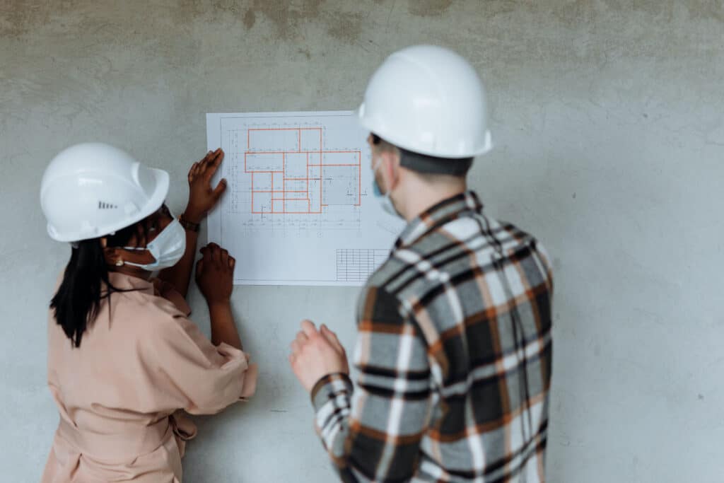 Two people wearing hard hats and masks discussing a floor plan on a wall.