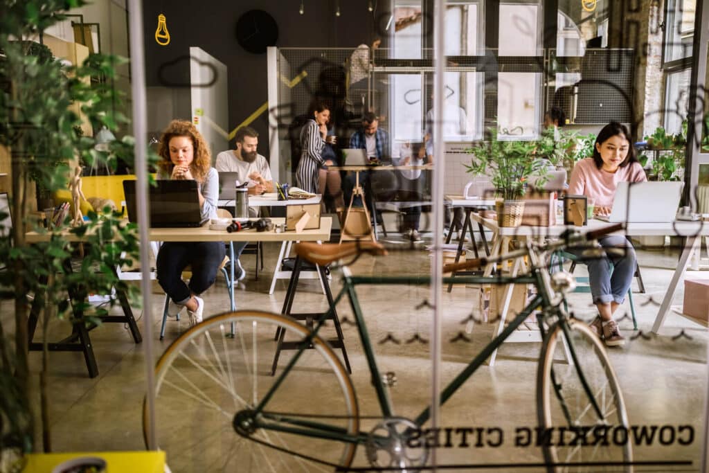 People working on laptops in a modern coworking space with plants and a bicycle in the foreground.