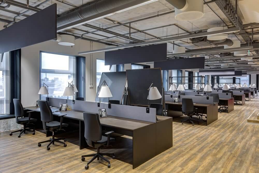 Modern open-plan office with multiple workstations, black chairs, and lamps, featuring large windows and wooden flooring.