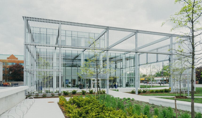 Modern building with a metal grid facade, surrounded by greenery and a cloudy sky.
