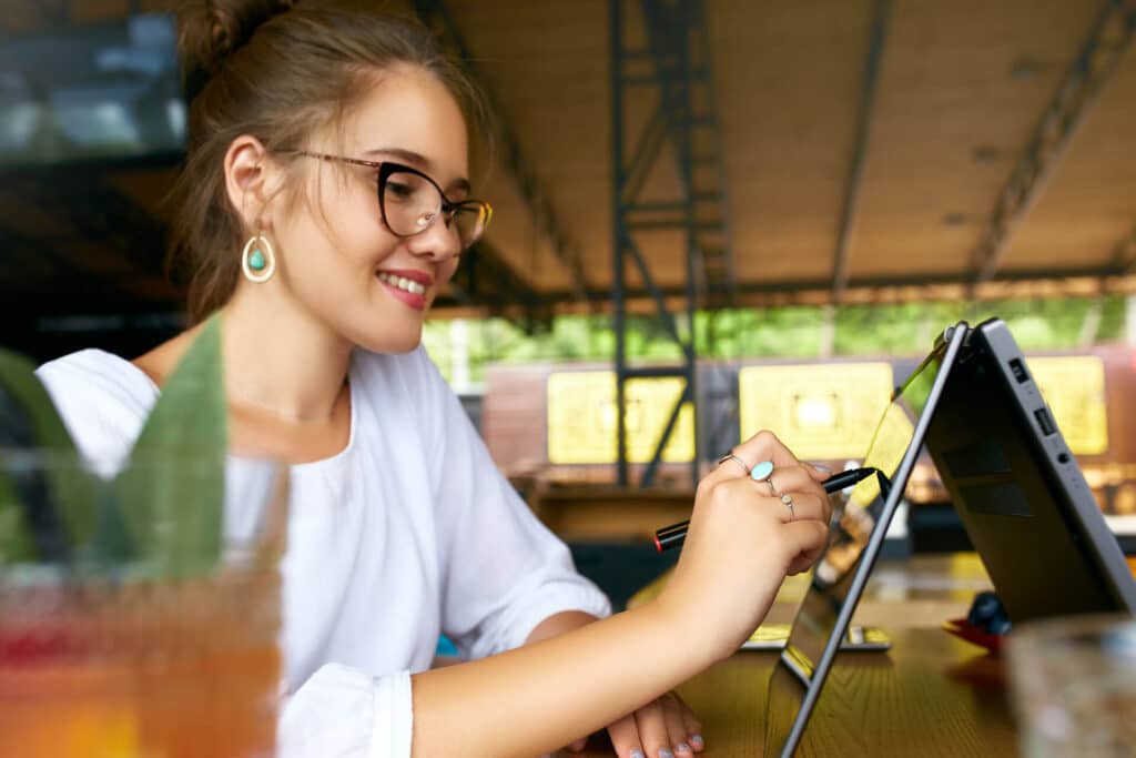 Woman with glasses uses a stylus on a tablet in a café, smiling.
