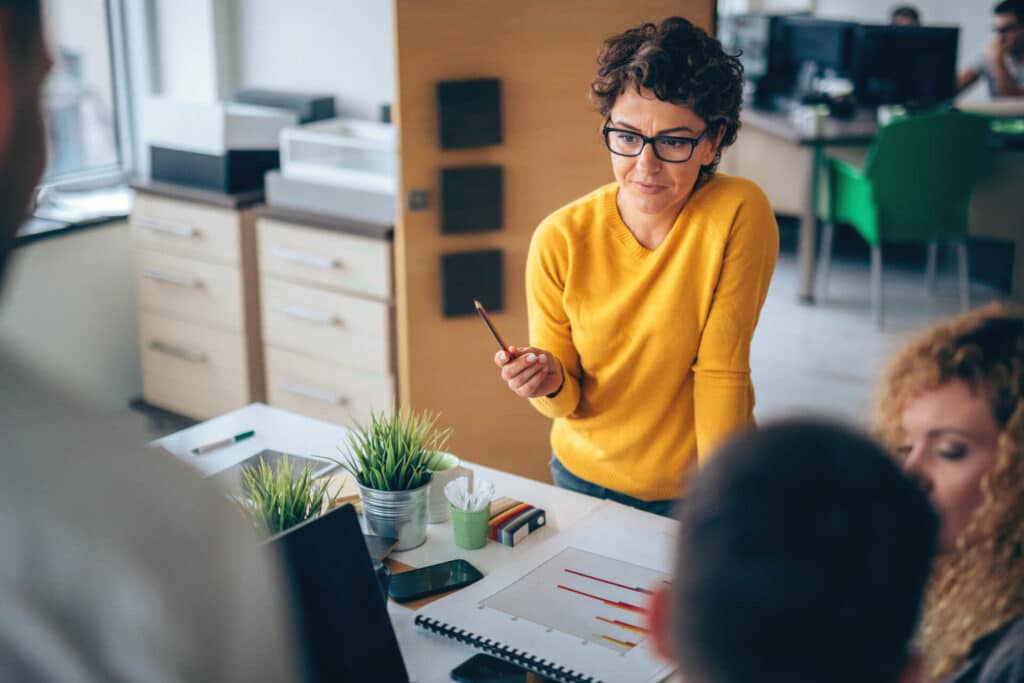 Woman in yellow sweater holds pen, discussing with colleagues around a table with charts and plants in office.