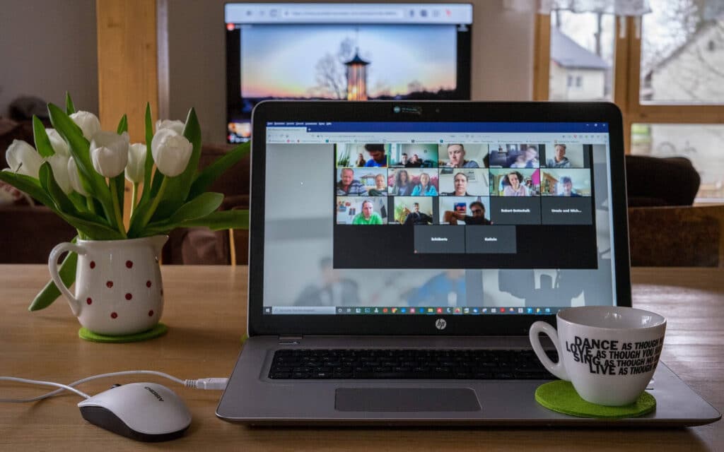 Laptop showing a video call with many participants, flanked by a flower vase and a coffee cup on a wooden desk.