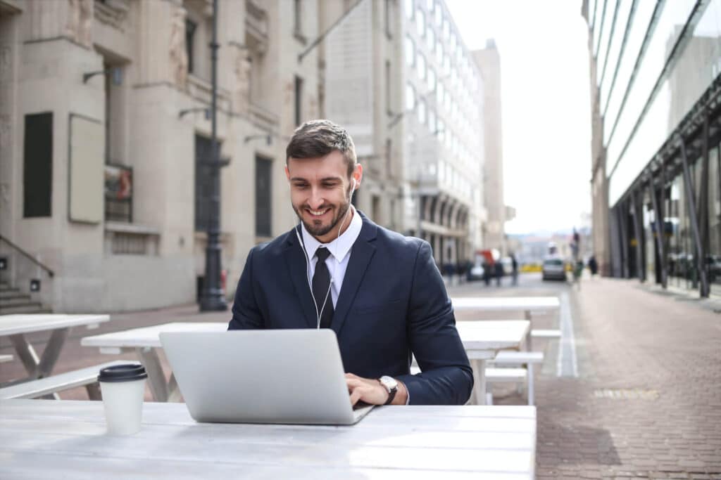 Man in a suit using a laptop and wearing earphones at an outdoor table, with city buildings in the background.