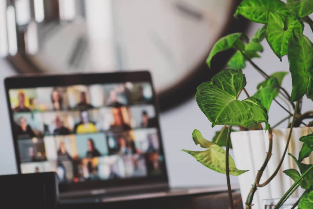 Laptop with a blurred video call on the screen, large wall clock behind, and a green plant on the right.