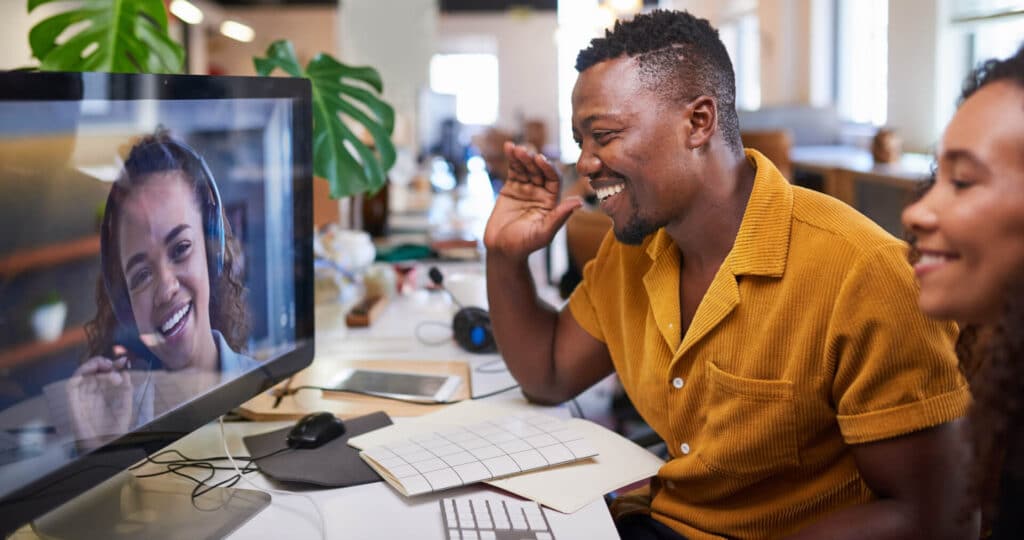 Man in yellow shirt and woman video call with a smiling person on a desktop screen in a modern office setting.