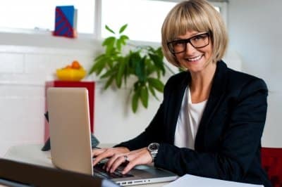 Person with glasses smiling while typing on a laptop at a desk with a plant and colorful decor in the background.