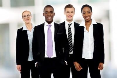 Four people in business attire standing together, smiling, with a blurred office background.