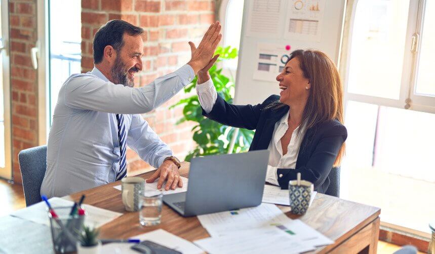 A man and woman in business attire high-five at a wooden table with a laptop, papers, and a mug.