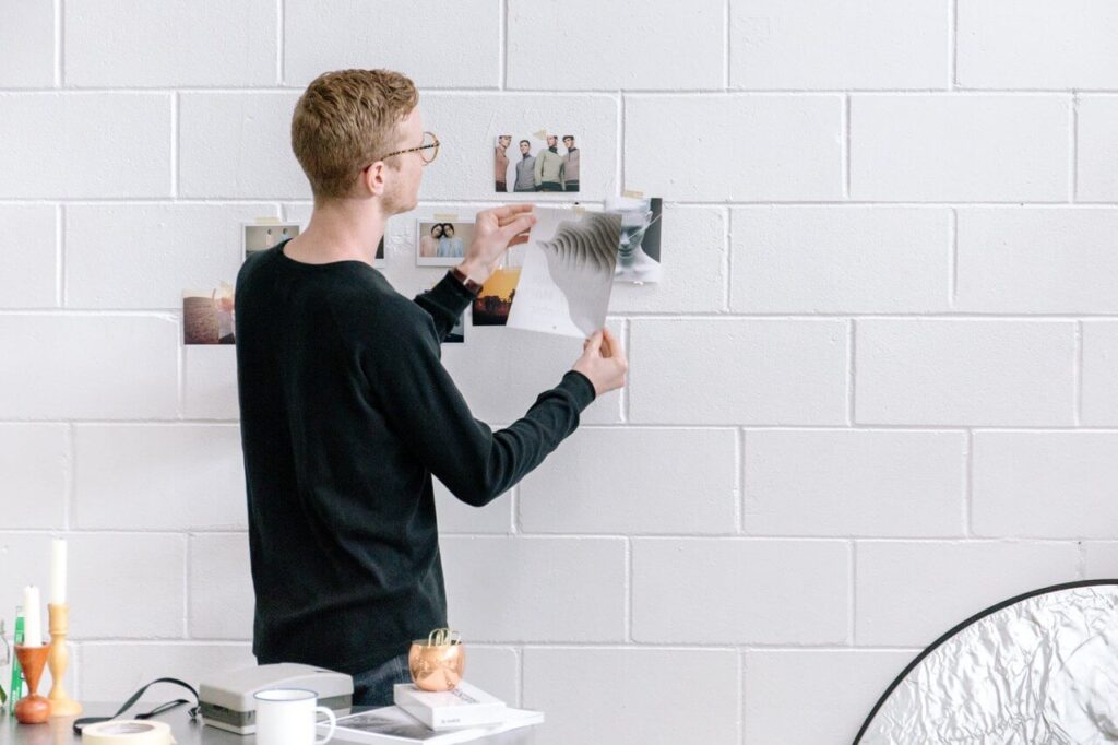 Man with glasses, in a black shirt, pinning photos to a white brick wall in a bright, modern room.