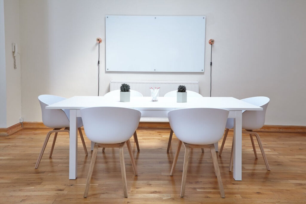 Minimalist meeting room with a white table, six white chairs, and a wall-mounted whiteboard in the background.