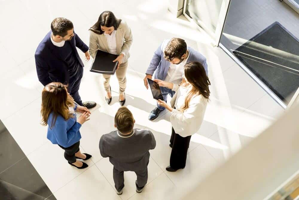 Group of six people standing and having a discussion in a brightly lit office space, seen from above.