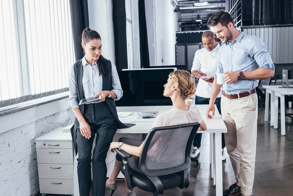 Four colleagues chatting and working in a modern office with computers and large windows.