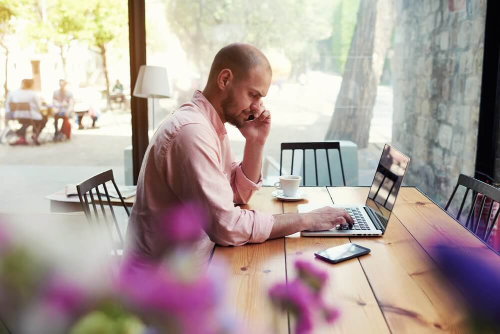 Man in pink shirt working on a laptop at a cafe, with a coffee cup and phone on the table, flowers in the foreground.