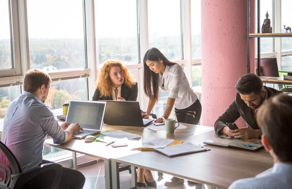 Group of people working around a table with laptops in a bright office setting.