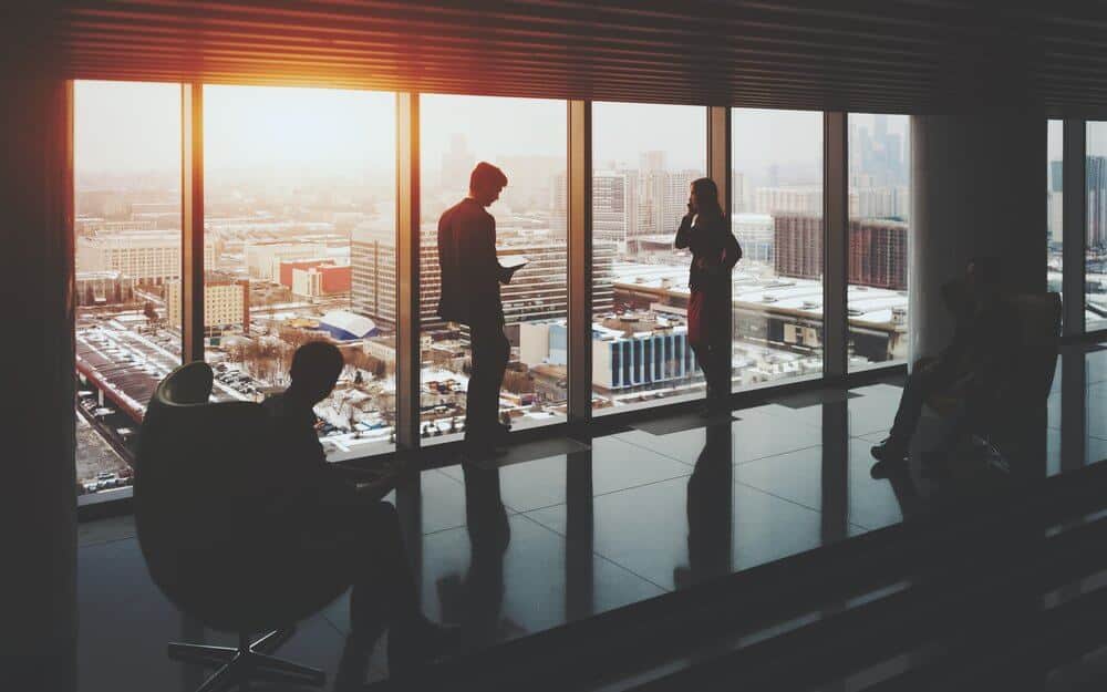 Silhouettes of people in a high-rise building, with a cityscape and sunset visible through large windows.