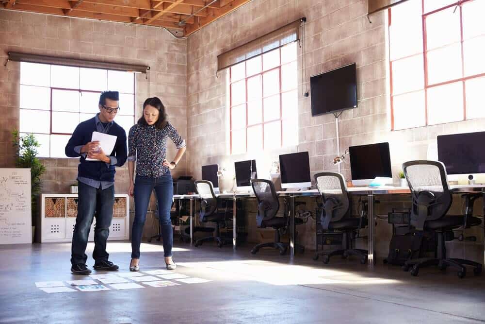 Two people review documents on the floor in a modern office with large windows and several workstations.