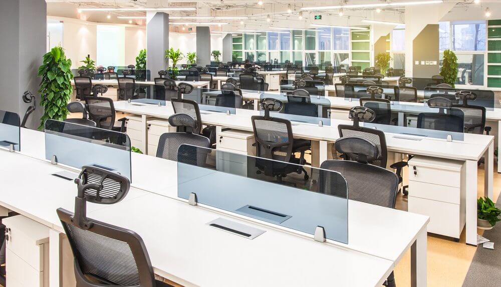 Modern open-plan office with rows of white desks, black chairs, and green plants under bright ceiling lights.