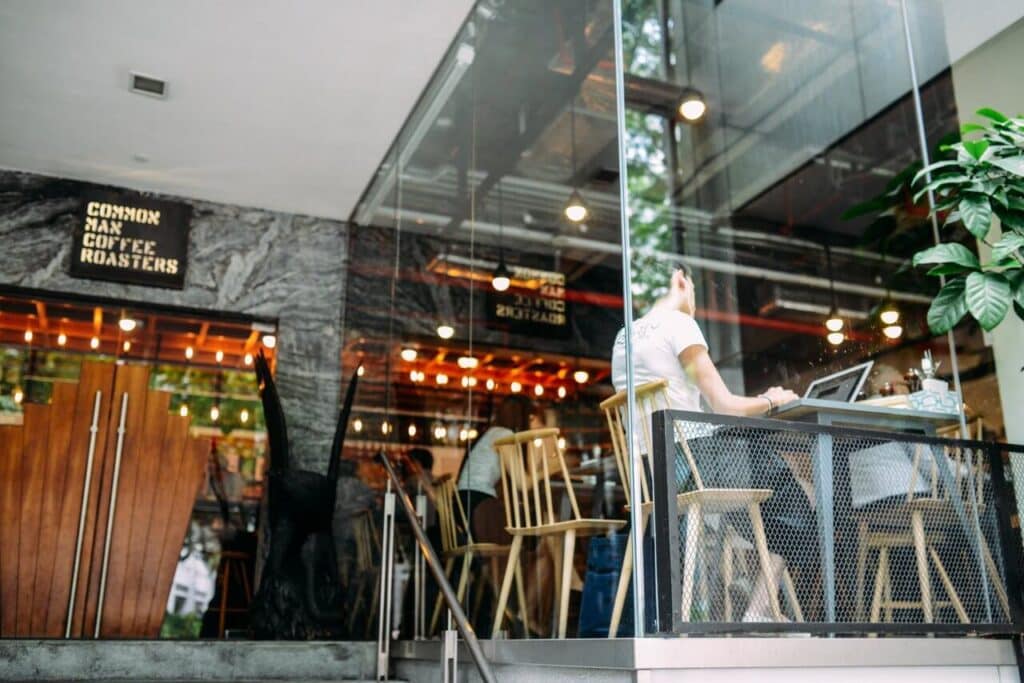 Person working on a laptop at an outdoor cafe with glass walls and wooden interior decor.