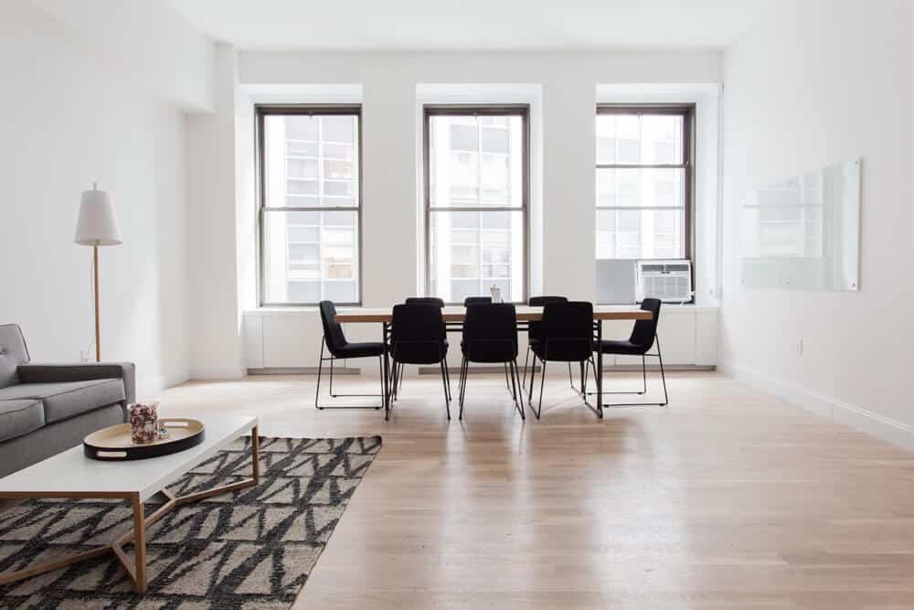 Minimalist room with a long table, black chairs, sofa, geometric rug, and three large windows.