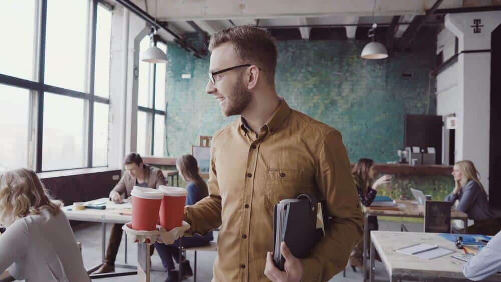 Man holding coffee cups and tablet in a modern office with colleagues working at desks in the background.