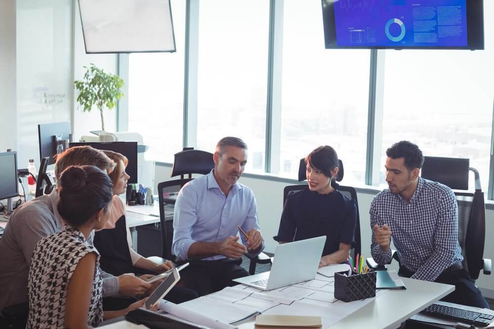 A group of professionals sitting around a table in an office, discussing and using a laptop.