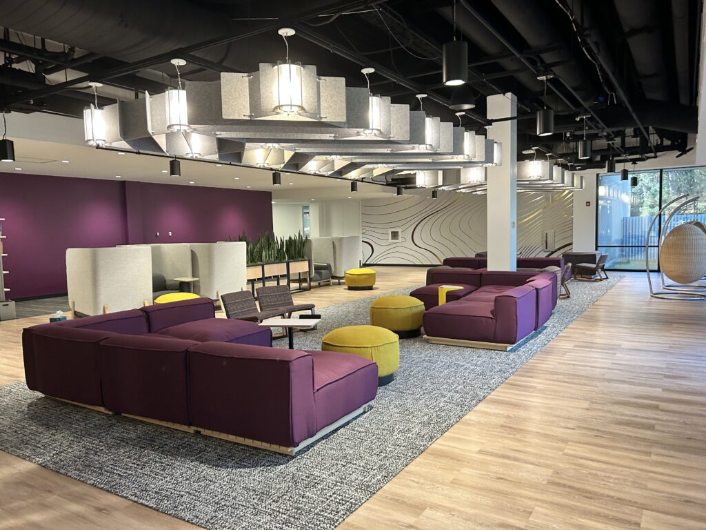 Modern lounge area with purple sofas, yellow ottomans, hanging lights, and geometric wall art.