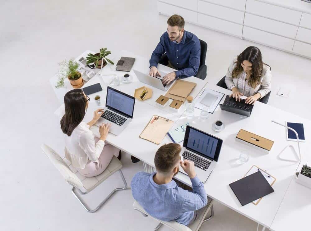 Top view of four people working on laptops at a white table in a bright office, with plants and office supplies.