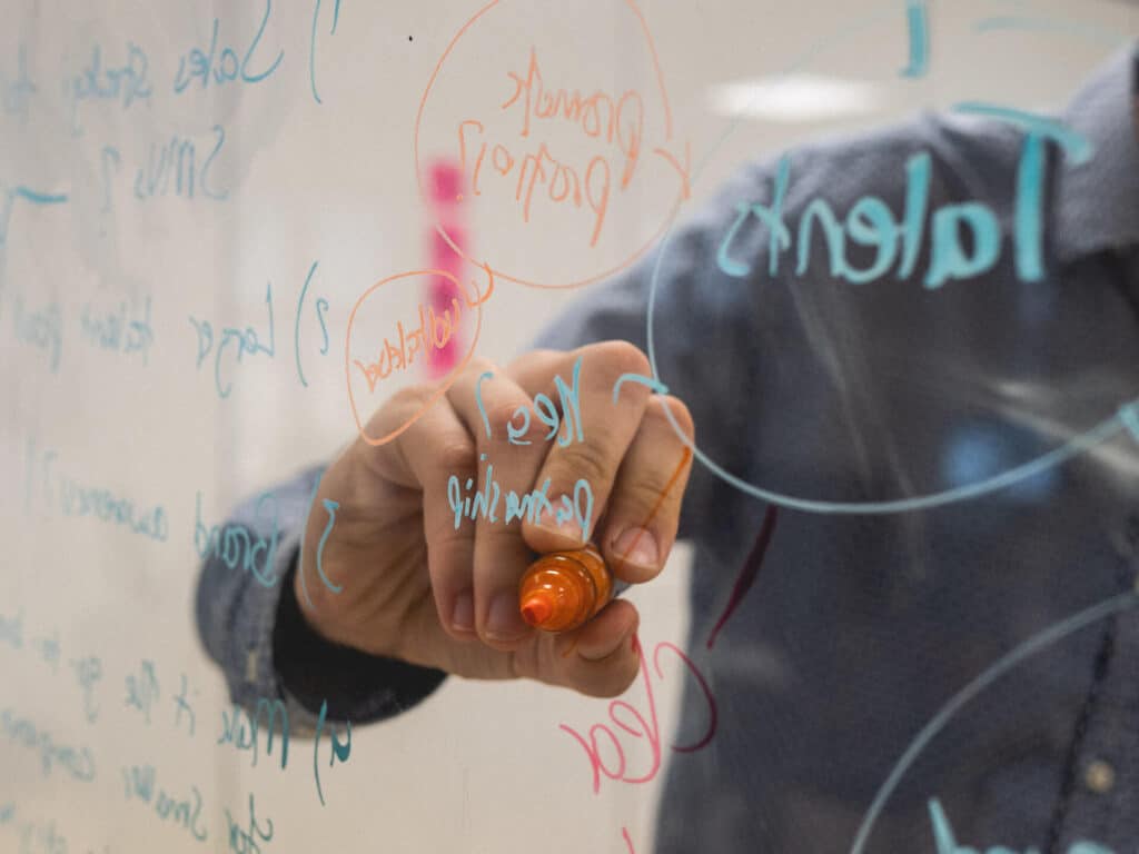 Person writing with orange marker on transparent board filled with various handwritten notes and diagrams.