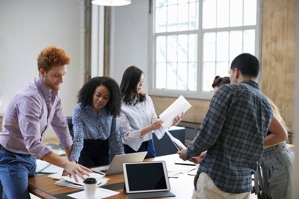 A group of people collaborates around a table with laptops and papers in a bright office.