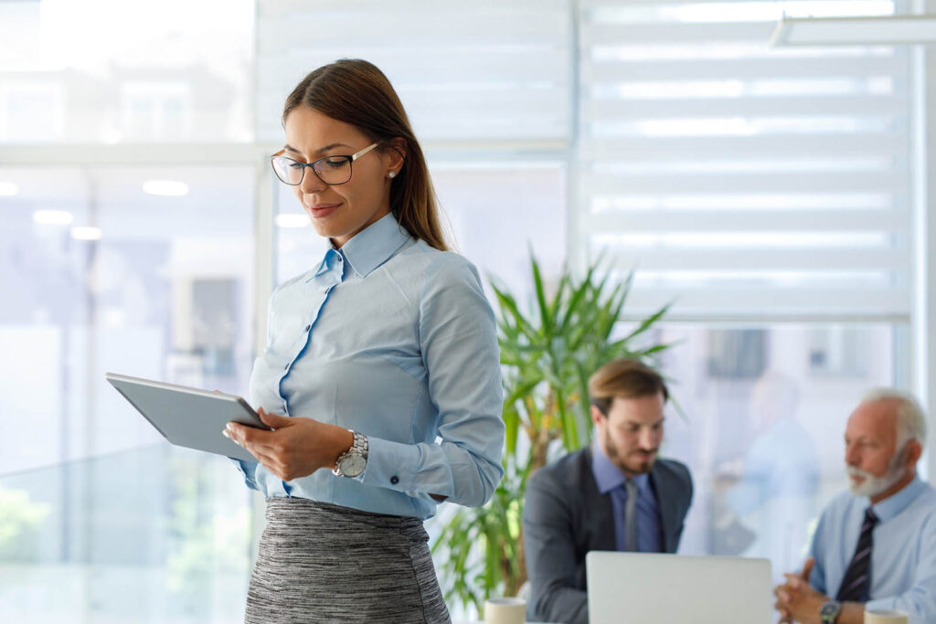 Businesswoman with tablet stands in an office; two men in suits work in the background.