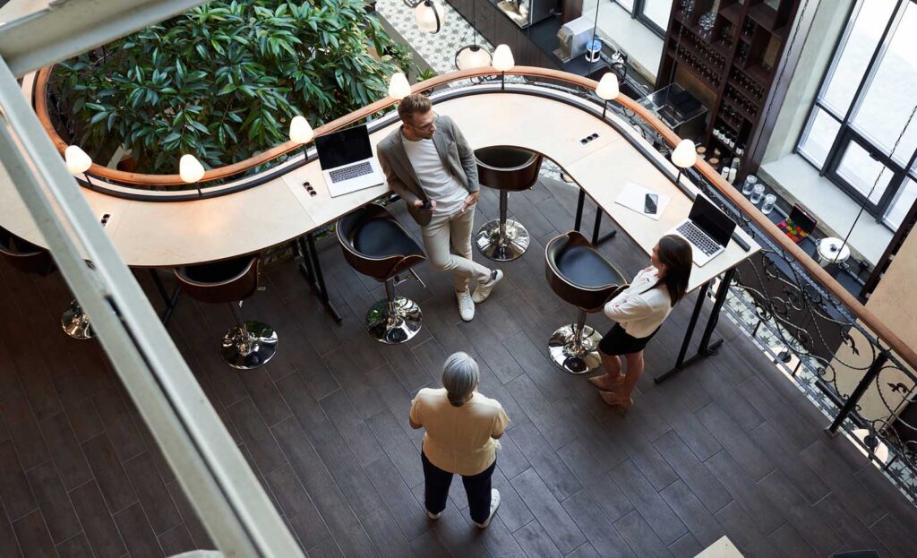 Three people have a meeting in a modern open-plan office with laptops on a curved desk, seen from above.