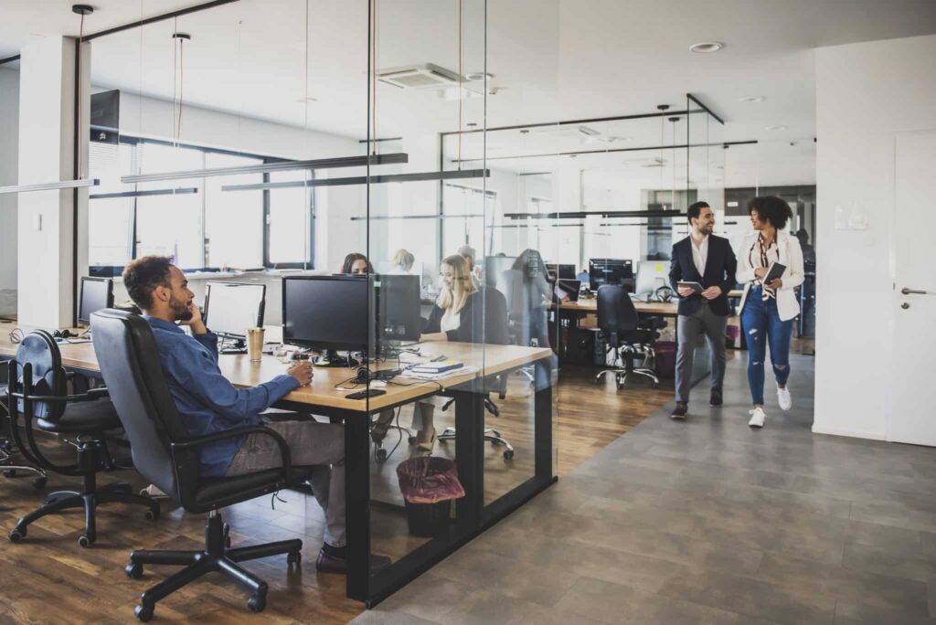 Open office space with people working at desks and two people walking and talking near the glass partition.