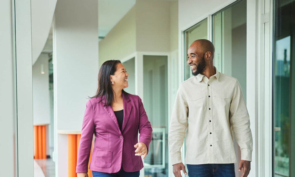 Two people walking and smiling in a bright hallway, with glass windows on one side.