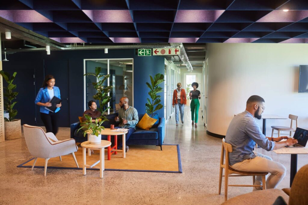 Open office with people working, plants, and modern décor. Some are seated, others walk and chat in the background.