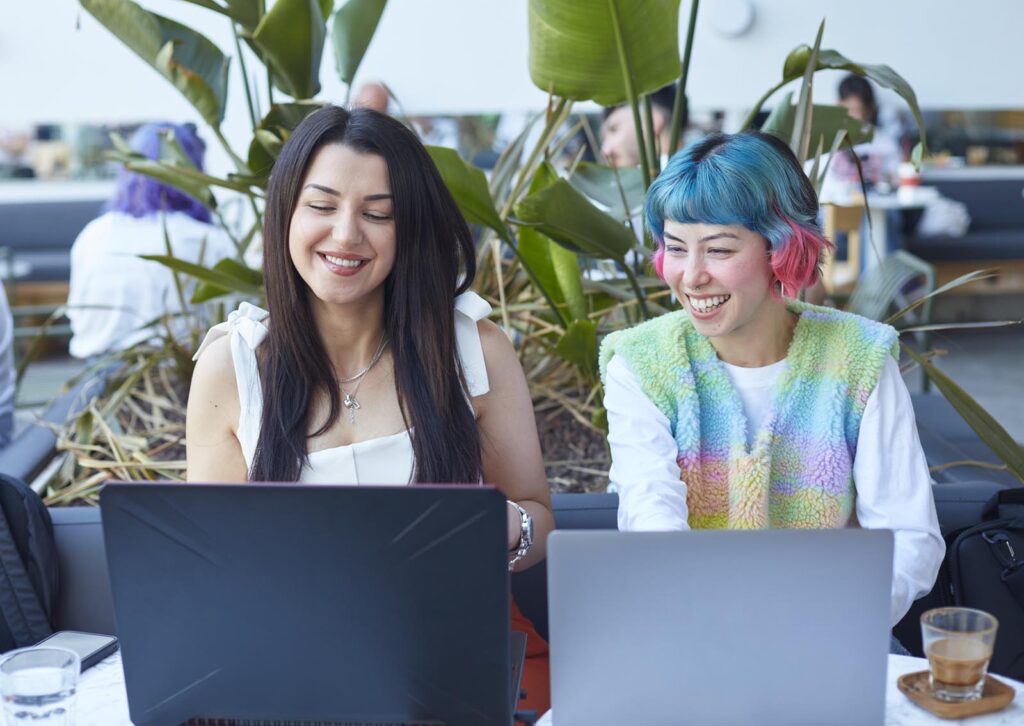 Two women smiling while working on laptops at a table with plants in the background.