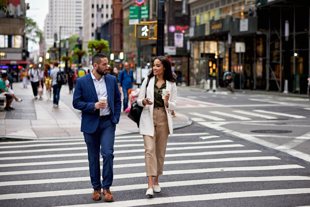 Full length front view of business associates in 20s and 30s crossing street, holding paper cups, talking and smiling.