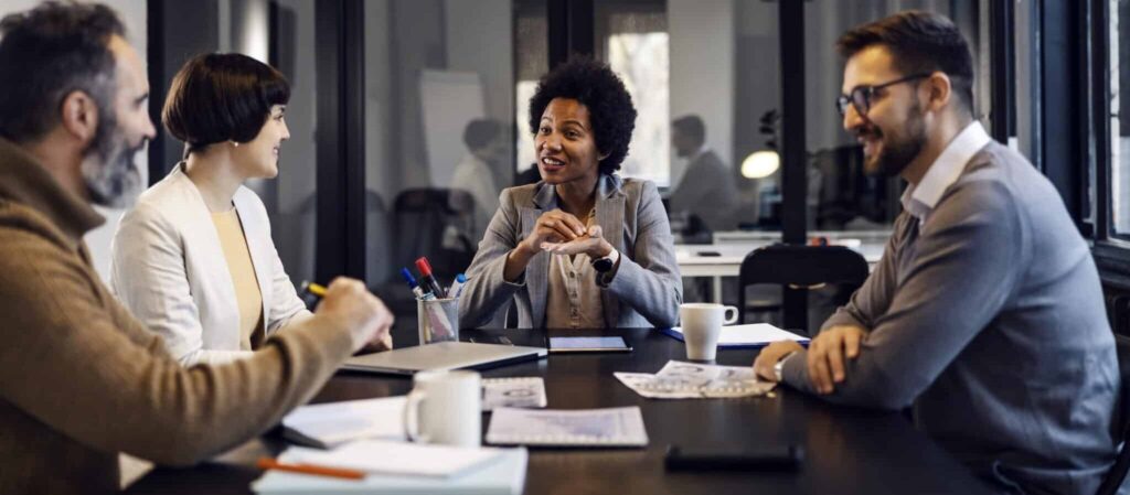 Four people in a meeting room engaged in a discussion, sitting around a table with papers and notebooks.