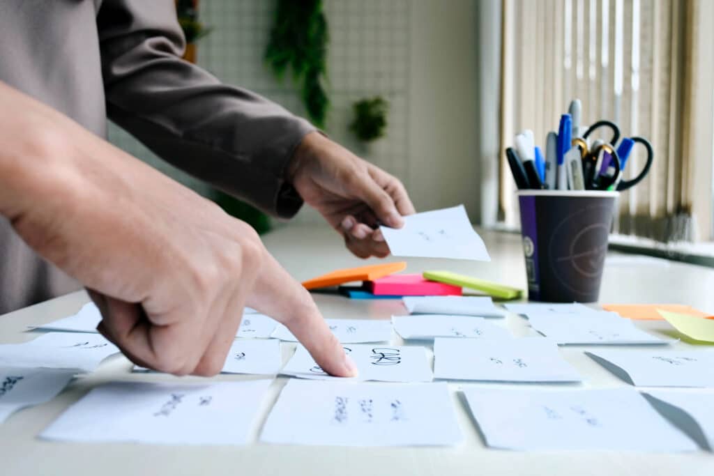 Hands organizing sticky notes on a table, with pens and a cup nearby.