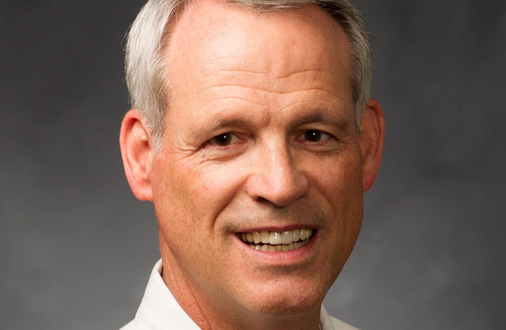 Smiling man with short gray hair wearing a white shirt against a gray background.