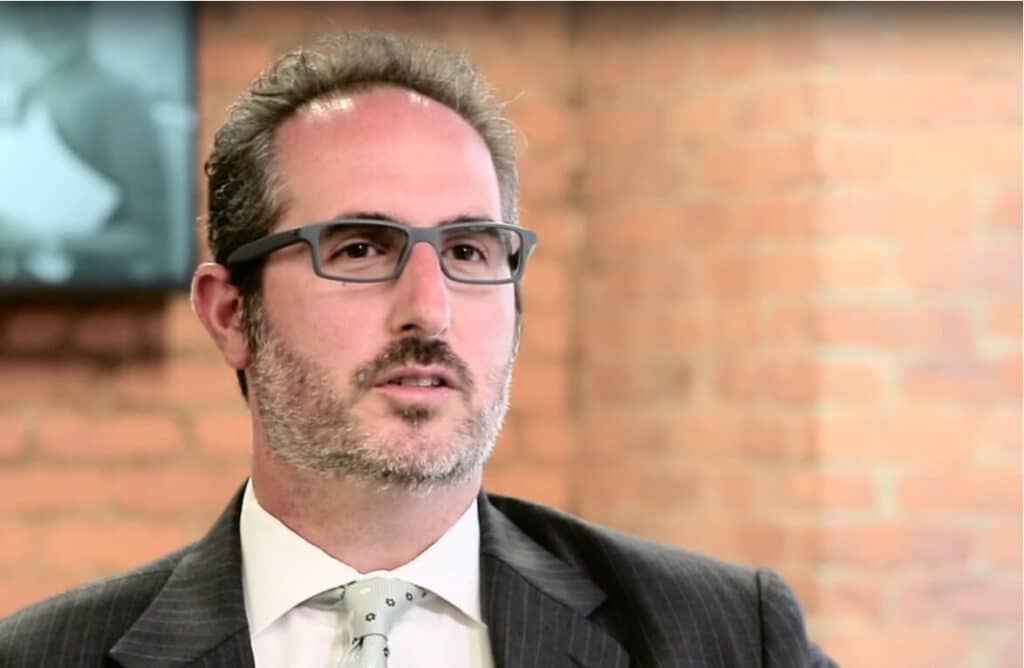 Man with glasses and beard in a suit, speaking in front of a brick wall background.