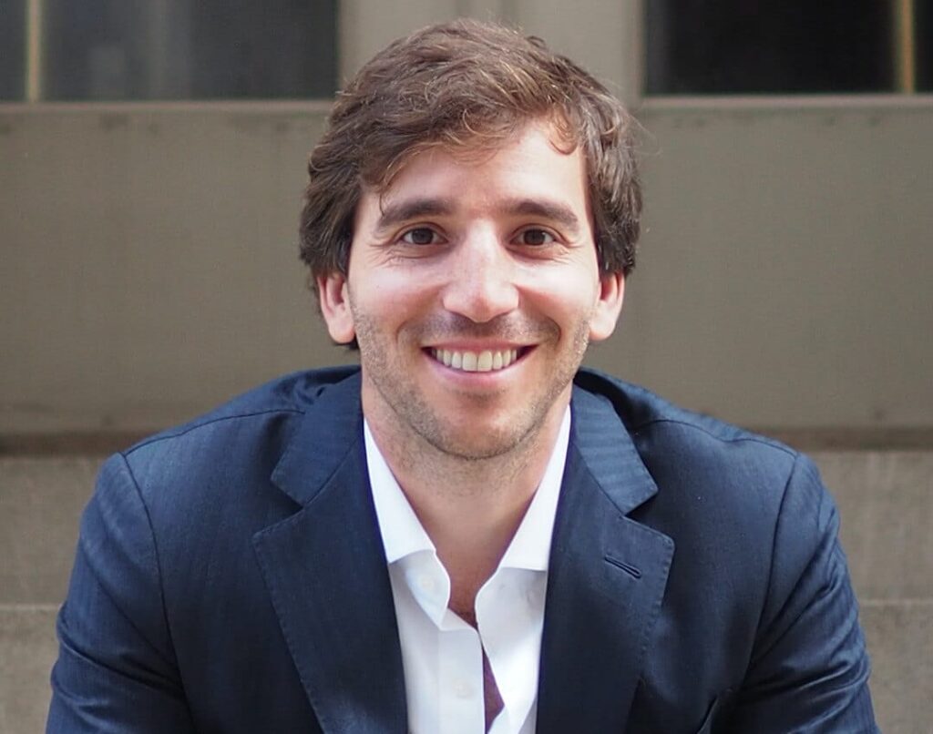 Smiling man in a dark suit and white shirt sitting in front of a building.