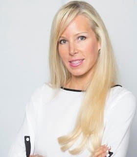 Woman with long blonde hair in a white blouse, arms crossed, smiling in front of a plain background.