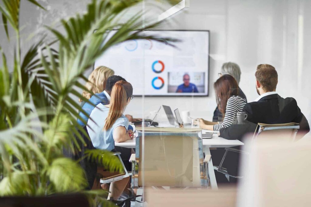 People in a conference room having a meeting with a video call on the screen, surrounded by glass walls and plants.