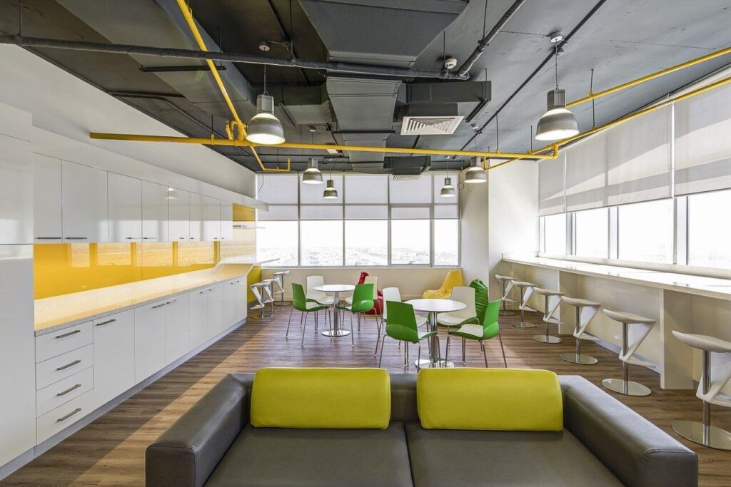 Modern office break room with a gray sofa, yellow and green chairs, round tables, and counter seating by large windows.